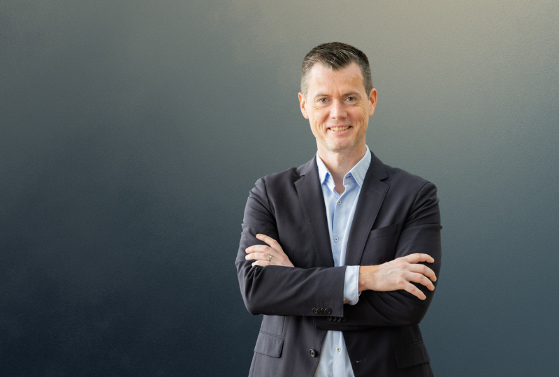 Person wearing a dark suit jacket over a light-colored dress shirt, standing with arms crossed against a gradient background